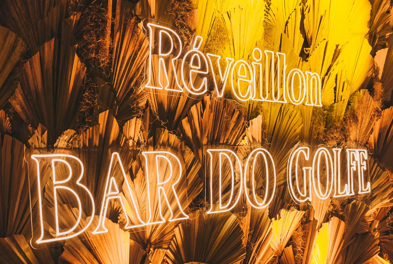 Letreiro em neon com o nome Réveillon Bar do Golfe sobre painel de folhas secas